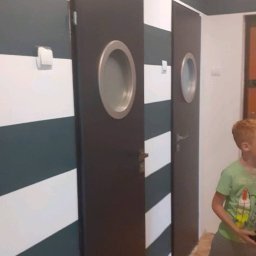 Remdambud - Pomalowane w poziome pasy ściany z dwoma ciemnymi drzwiami z okrągłymi okienkami. Widoczny grzejnik i fragment osoby w pomieszczeniu.