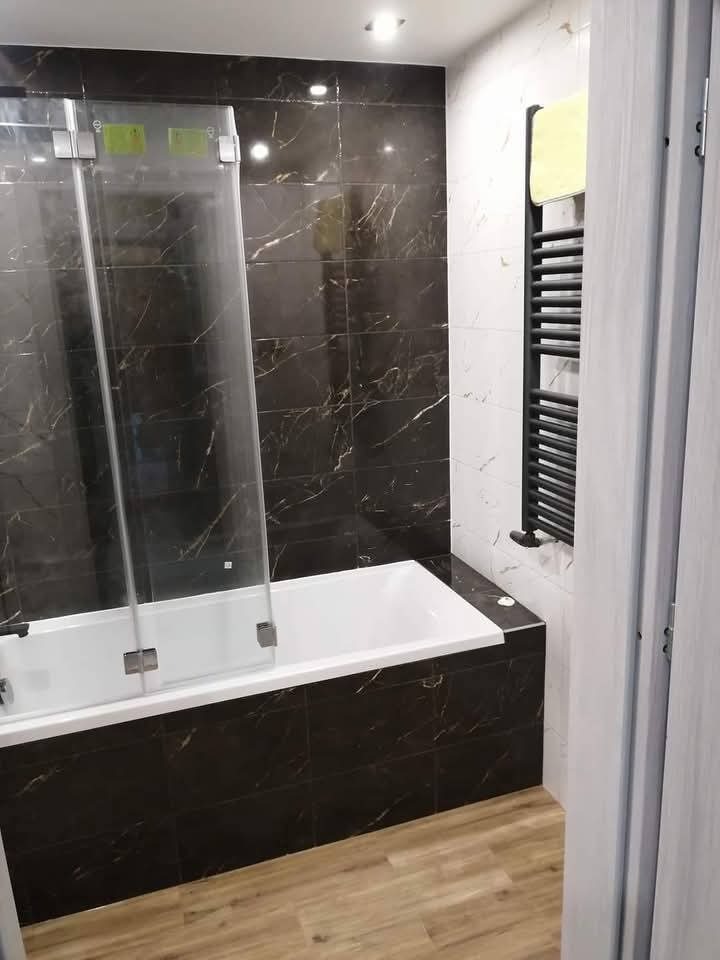 Wykończona łazienka z wanną obudowaną czarnymi płytkami imitującymi marmur, szklanym parawanem, grzejnikiem drabinkowym i podłogą imitującą drewno.
