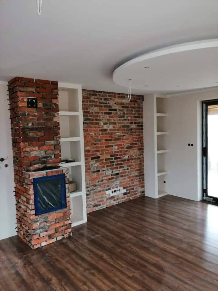 Wykończone wnętrze z ceglaną ścianą i kominkiem, drewniana podłoga, białe półki w ścianie. Nowoczesny design z elementami rustykalnymi.