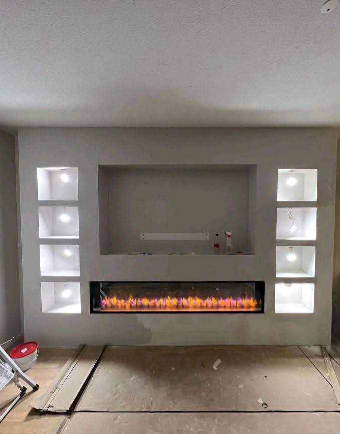 Ściana z wnękami i elektrycznym kominkiem w trakcie wykończenia. Widoczne kartony ochronne na podłodze i drabina. Górne wnęki podświetlone punktowo.