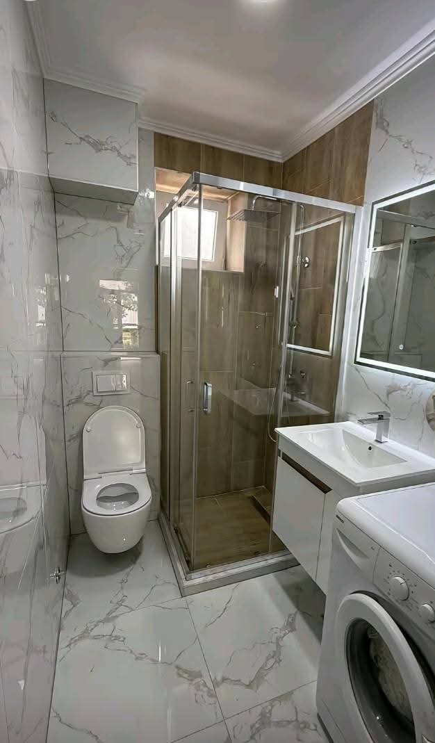 Wykończona łazienka w Warszawie: marmurowe płytki, kabina prysznicowa z jasnymi drzwiami, toaleta i pralka. Nowoczesny design z oświetleniem LED przy lustrze.