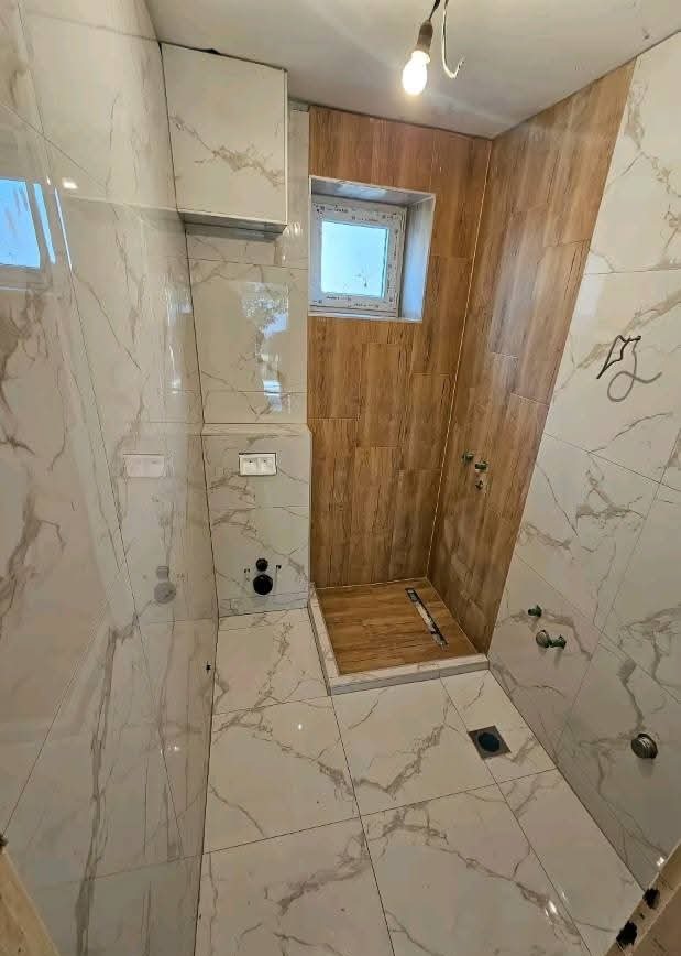 Wykończona łazienka z kabiną prysznicową wyłożoną drewnopodobnymi płytkami. Na ścianach i podłodze marmurowe płytki. Widoczne elementy armatury i okno.