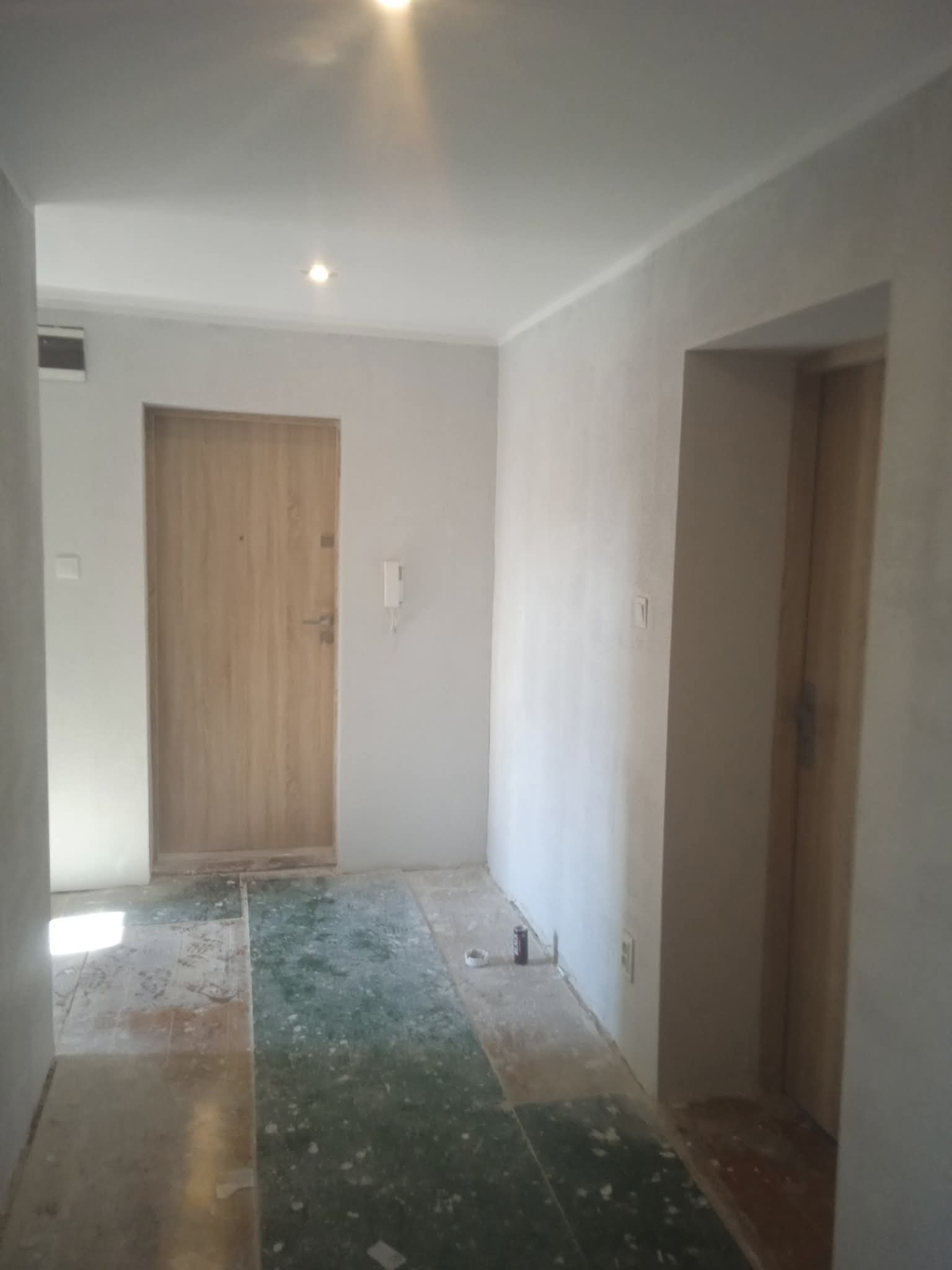 Wnętrze w trakcie remontu: szpachlowane ściany, widoczne ślady po starej podłodze, drzwi wejściowe. Etap przygotowania do dalszych prac wykończeniowych.