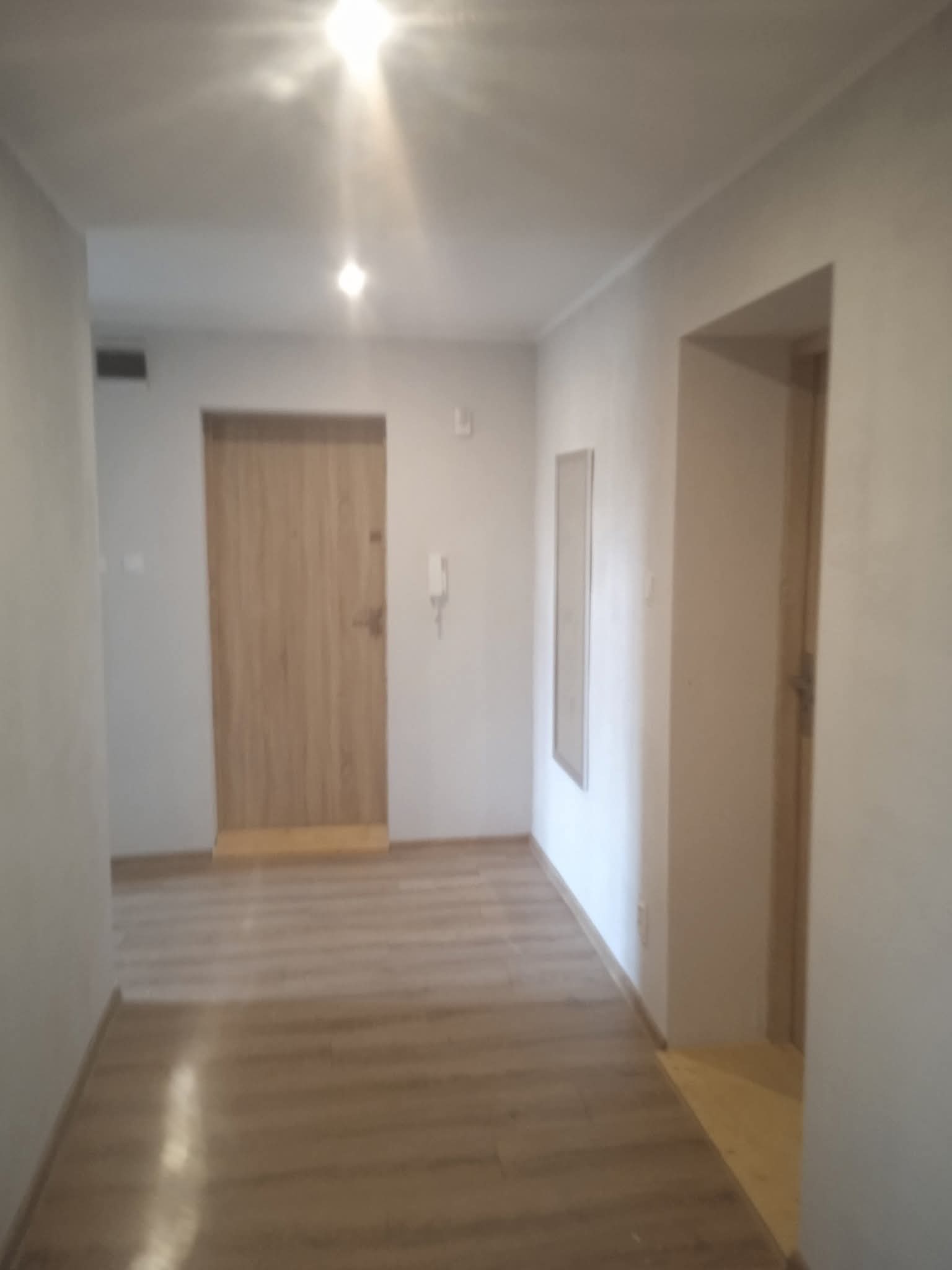 Korytarz po remoncie: odnowione ściany w jasnym odcieniu, nowe panele podłogowe imitujące drewno, drzwi wejściowe i wewnętrzne. Minimalistyczny design.
