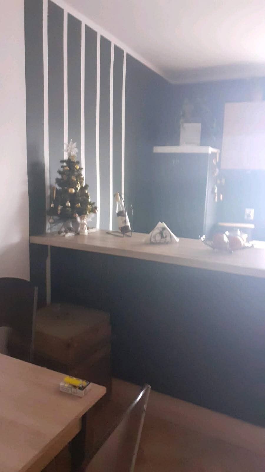 Szaro-biała ściana z pionowymi pasami za blatem, na którym stoi mała choinka. W tle widoczna kuchnia. U dołu fragment stołu i krzesła.