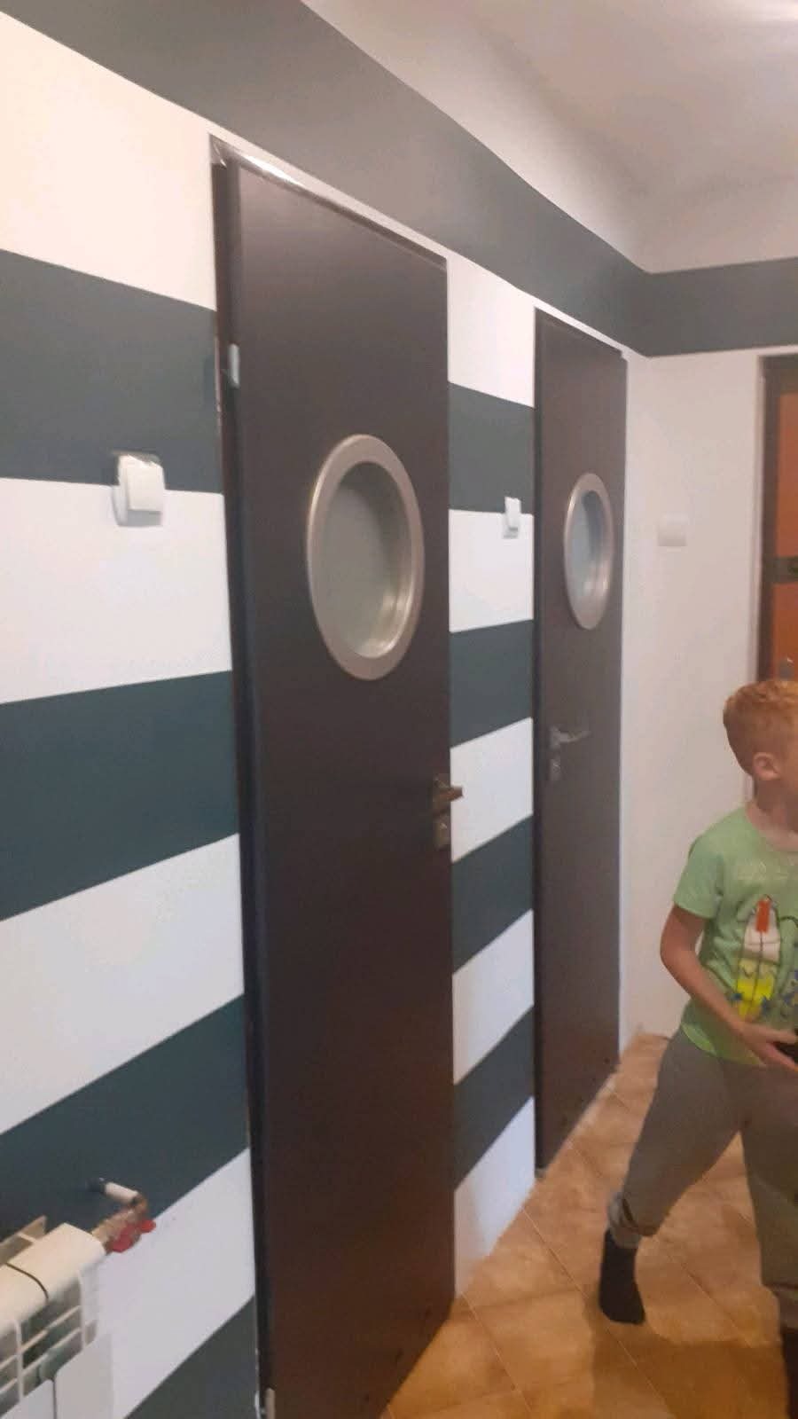 Pomalowane w poziome pasy ściany z dwoma ciemnymi drzwiami z okrągłymi okienkami. Widoczny grzejnik i fragment osoby w pomieszczeniu.