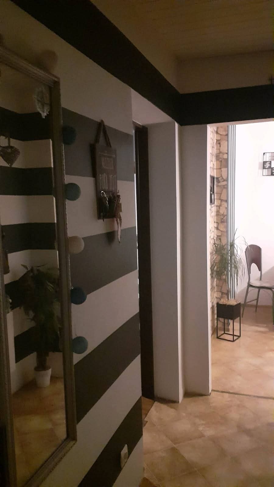 Wnętrze po malowaniu: ściana w biało-szare pasy, lustro, dekoracje. Widoczny fragment podłogi z płytek i dalsza część pomieszczenia z rośliną i krzesłem.