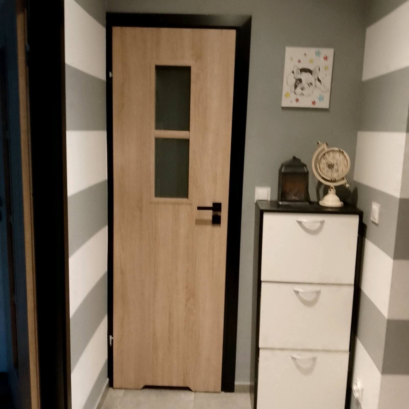 Drewniane drzwi z czarną ramą i przeszkleniami, zamontowane w pokoju z szaro-białymi pasami na ścianie. Obok komoda z dekoracjami.