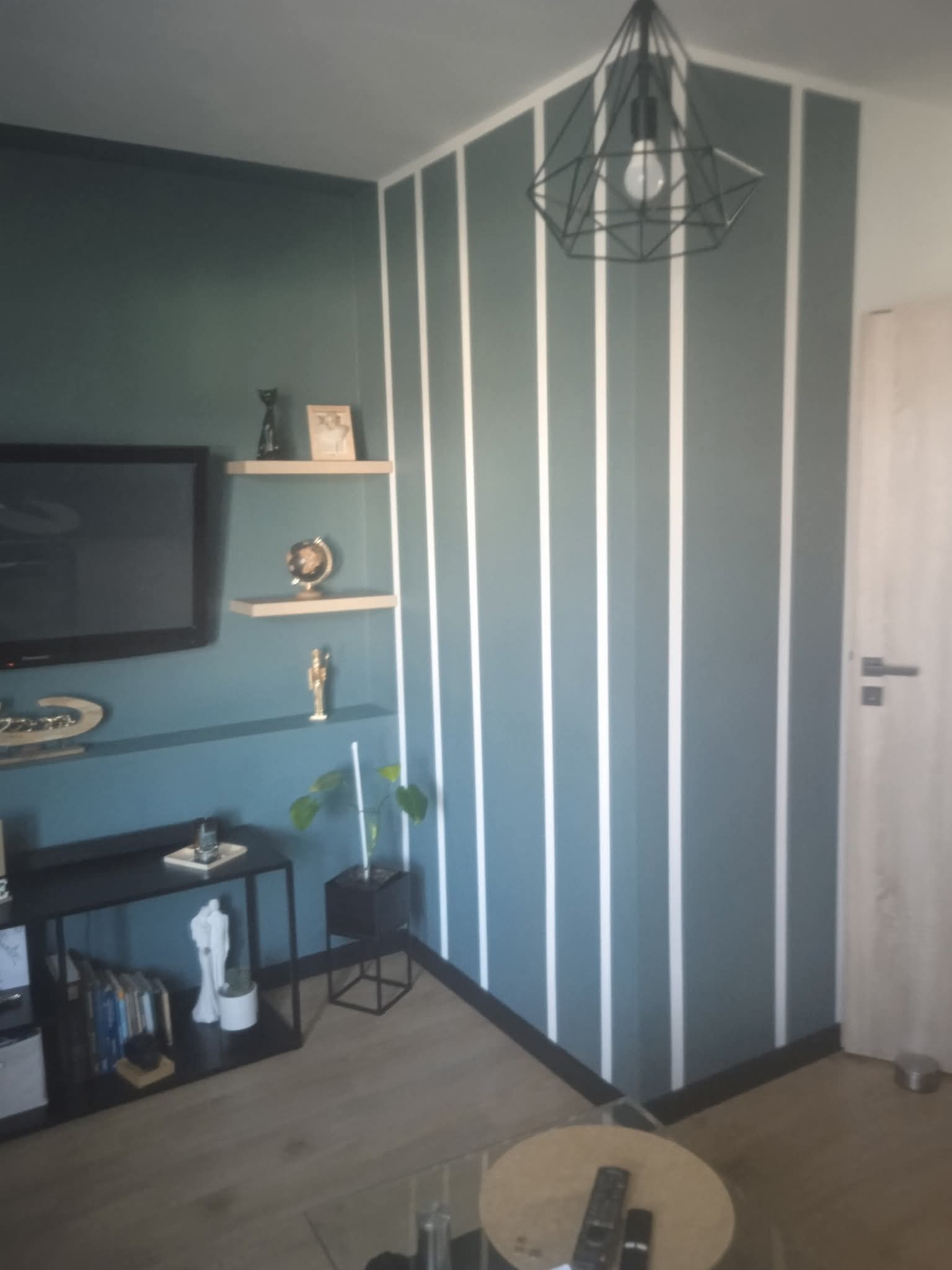 Niebieska ściana z białymi pasami w nowoczesnym salonie. Na ścianie wiszą półki z dekoracjami, a pod sufitem geometryczna lampa. Widoczny fragment telewizora i drzwi.