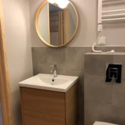 Nowoczesna łazienka z drewnianą szafką pod umywalką, okrągłym lustrem i grzejnikiem drabinkowym. Szare płytki i minimalistyczna armatura.