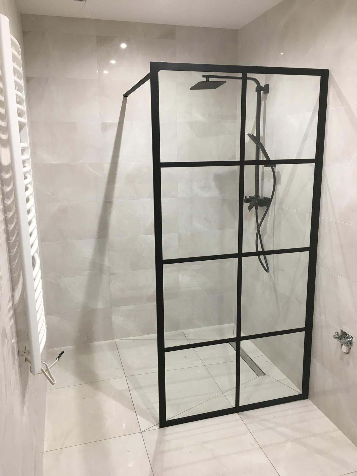 Nowoczesna kabina prysznicowa z czarną ramą i deszczownicą, jasne płytki, grzejnik drabinkowy. Minimalistyczny design łazienki po remoncie.