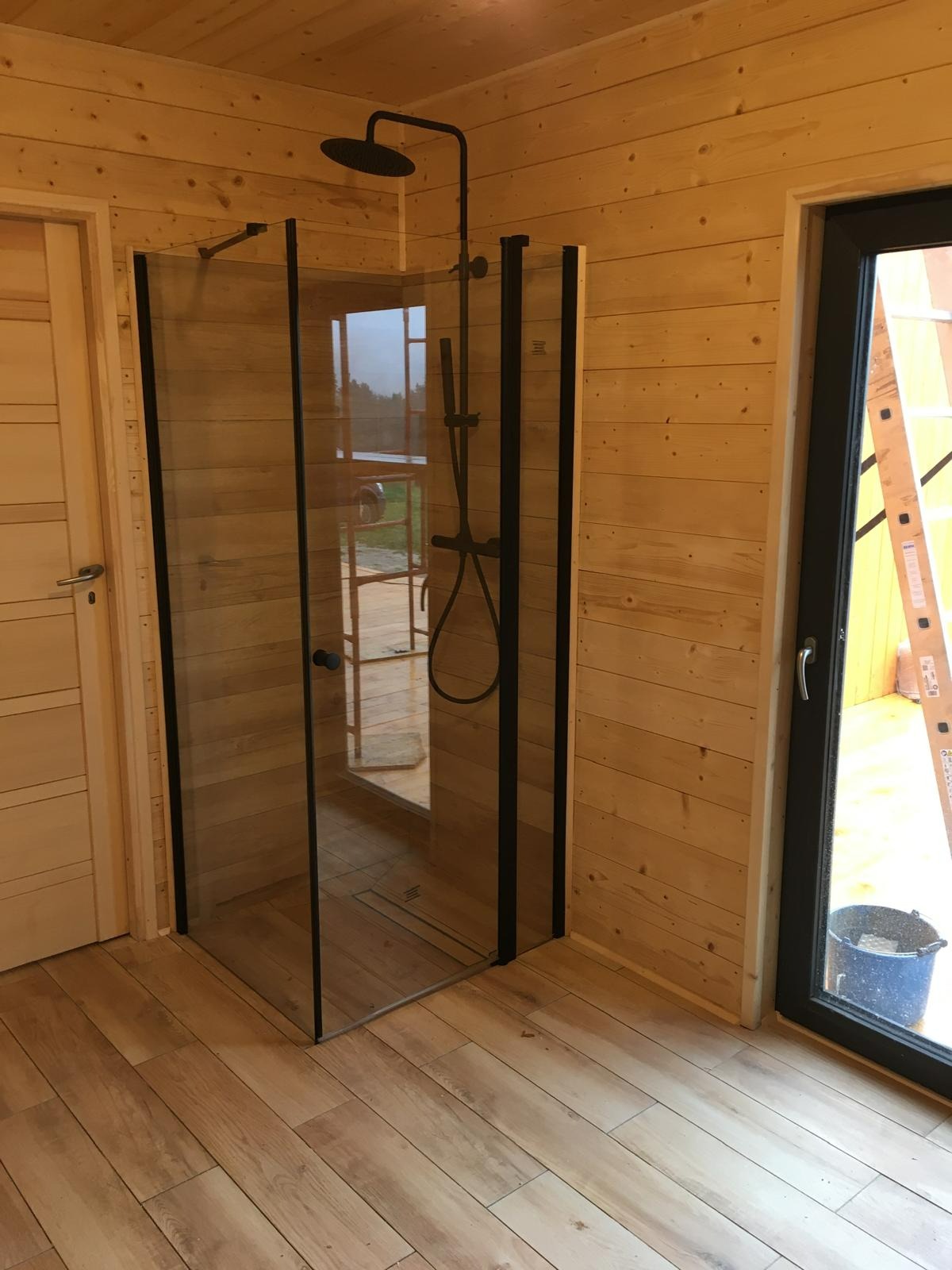 Kabina prysznicowa z czarnymi ramami i deszczownicą w drewnianej łazience. Widoczne drzwi i okno, podłoga wyłożona panelami imitującymi drewno.