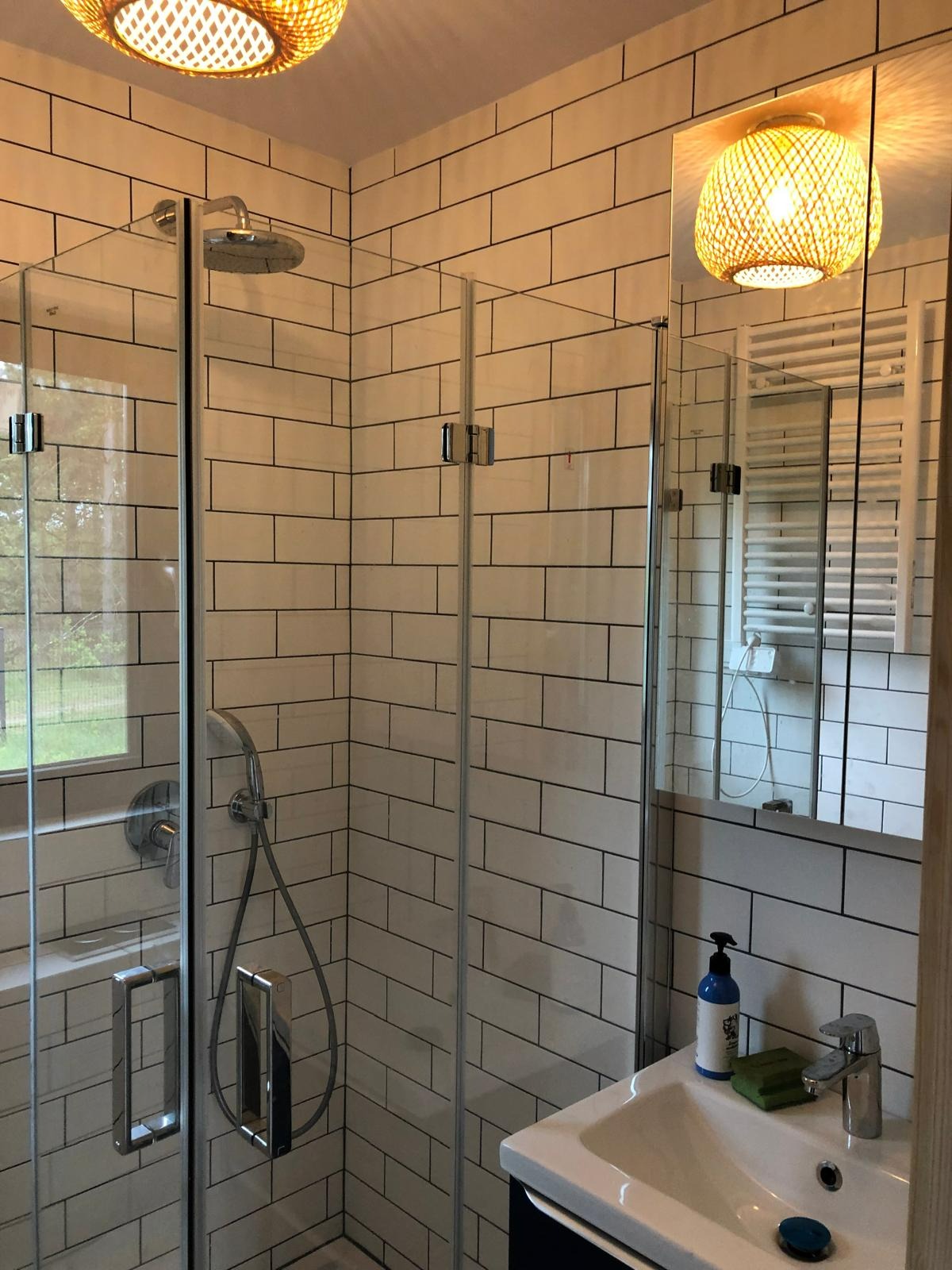 Biała łazienka z kabiną prysznicową typu walk-in, wykończona płytkami imitującymi cegłę, oświetlona dwoma plecionymi lampami, z umywalką i lustrem.