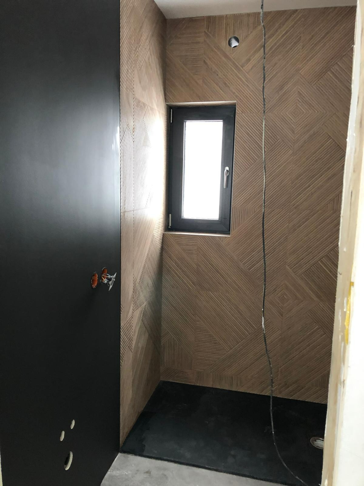 Wnętrze łazienki w trakcie wykańczania: wzorzyste płytki na ścianach, czarna kabina prysznicowa i okno z czarną ramą. Widoczne przewody elektryczne i otwory na gniazdka.