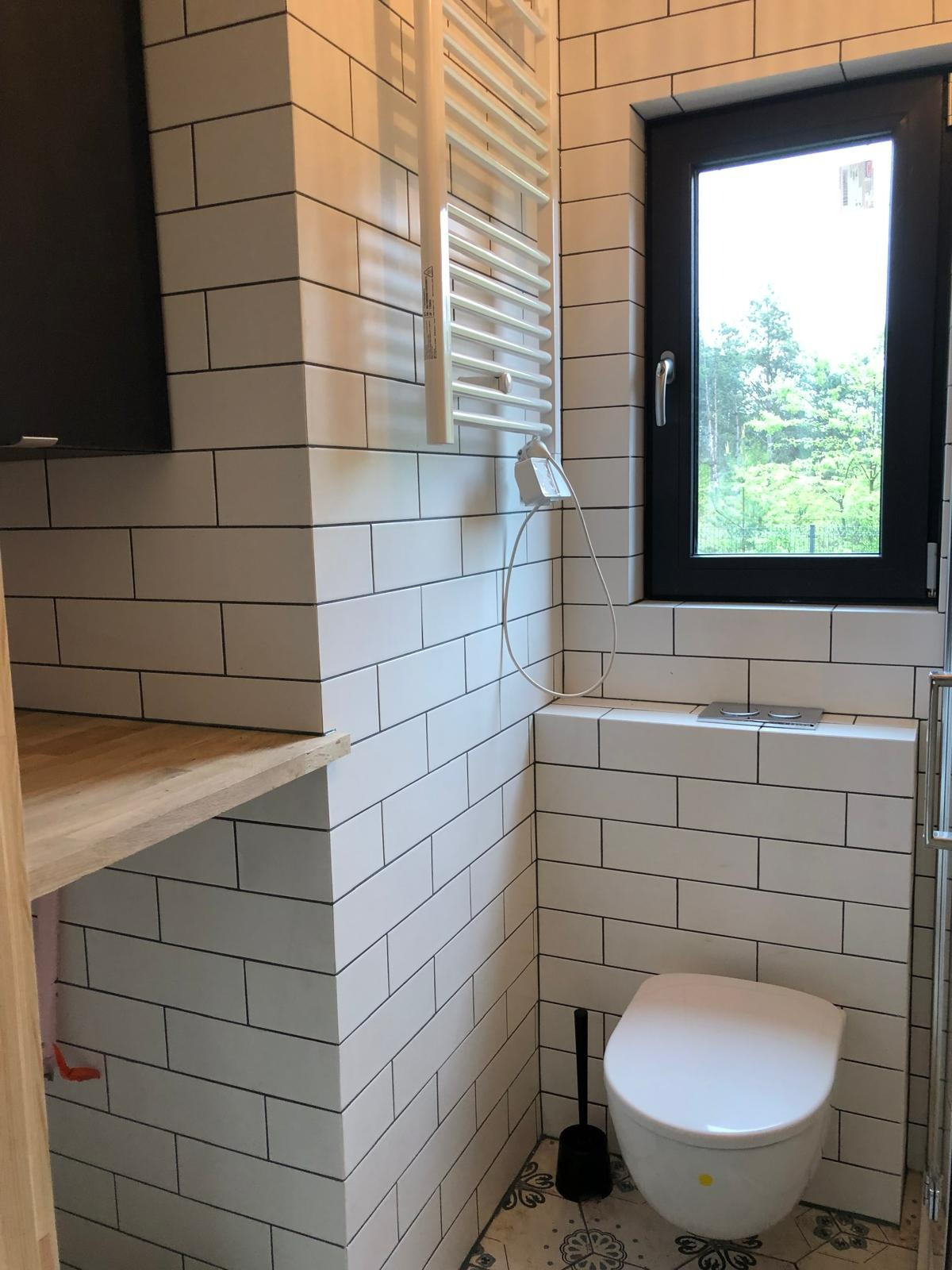 Nowoczesna łazienka po remoncie z białymi płytkami imitującymi cegłę, wiszącą toaletą i grzejnikiem drabinkowym. Widok na okno z zielenią w tle.