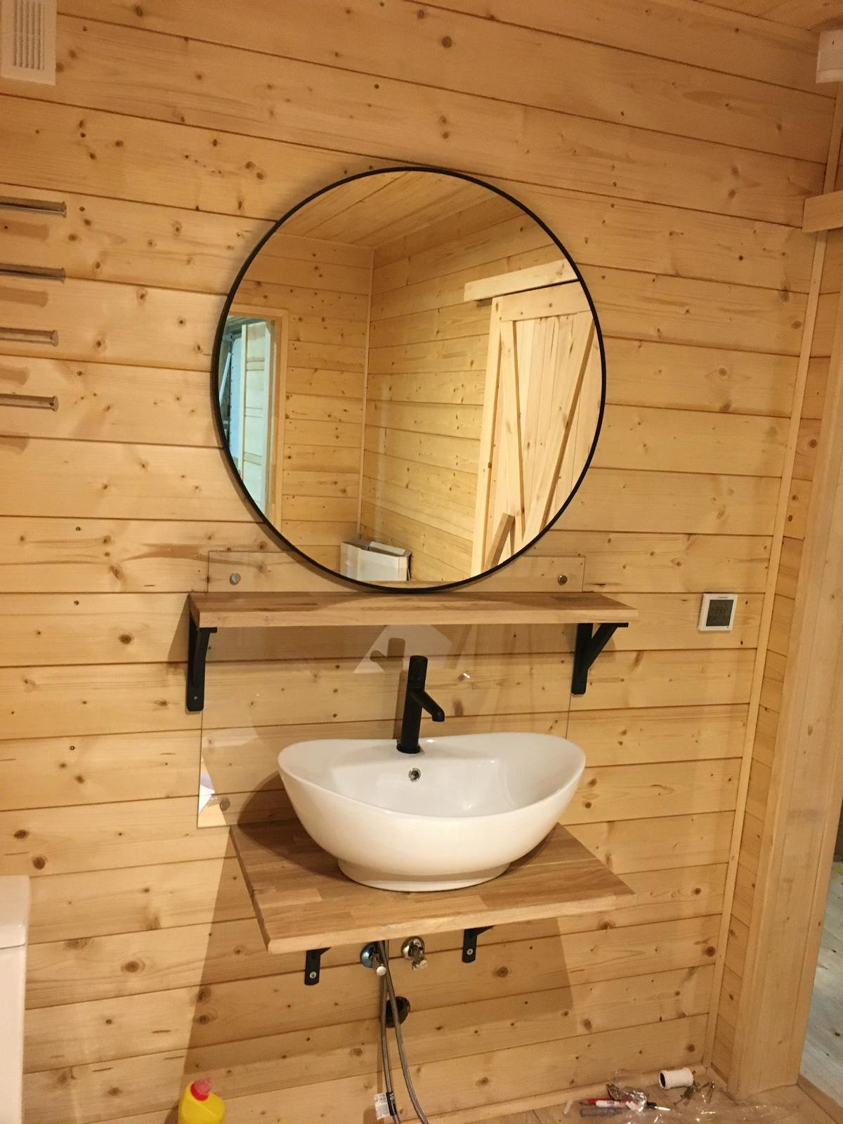 Nowoczesna umywalka nablatowa w białej misie, zamontowana na drewnianym blacie w łazience wykończonej boazerią. Okrągłe lustro z czarną ramą nad umywalką.