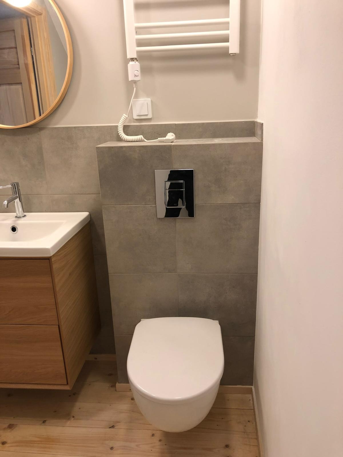Ukończona łazienka z drewnianą szafką pod umywalką, wiszącą toaletą i grzejnikiem drabinkowym. Szare płytki na ścianie, drewniana podłoga.