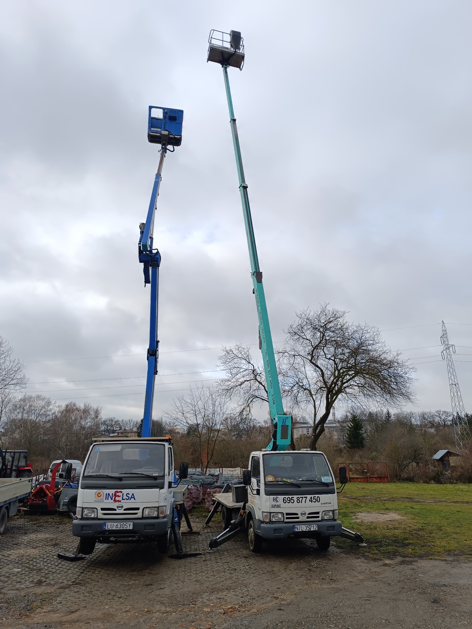 Dwa podnośniki koszowe na białych samochodach dostawczych, jeden niebieski, drugi zielony, stojące na placu pod pochmurnym niebem. Widoczne podpory stabilizujące.
