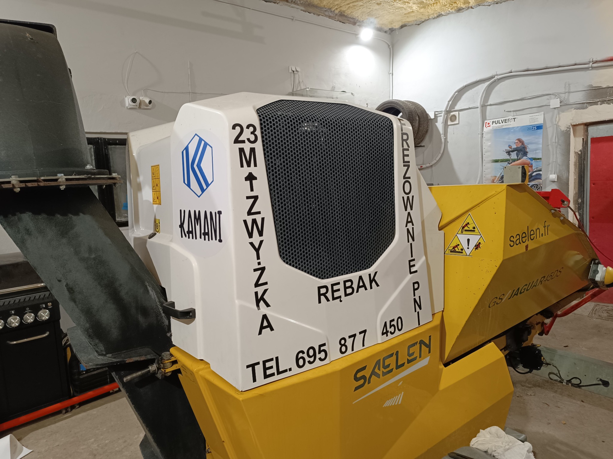 Żółto-biały rębak do gałęzi marki Saelen GS Jaguar 45DS z widocznym logo Kamani, numerem telefonu i napisem 'Frezowanie pni' w pomieszczeniu.