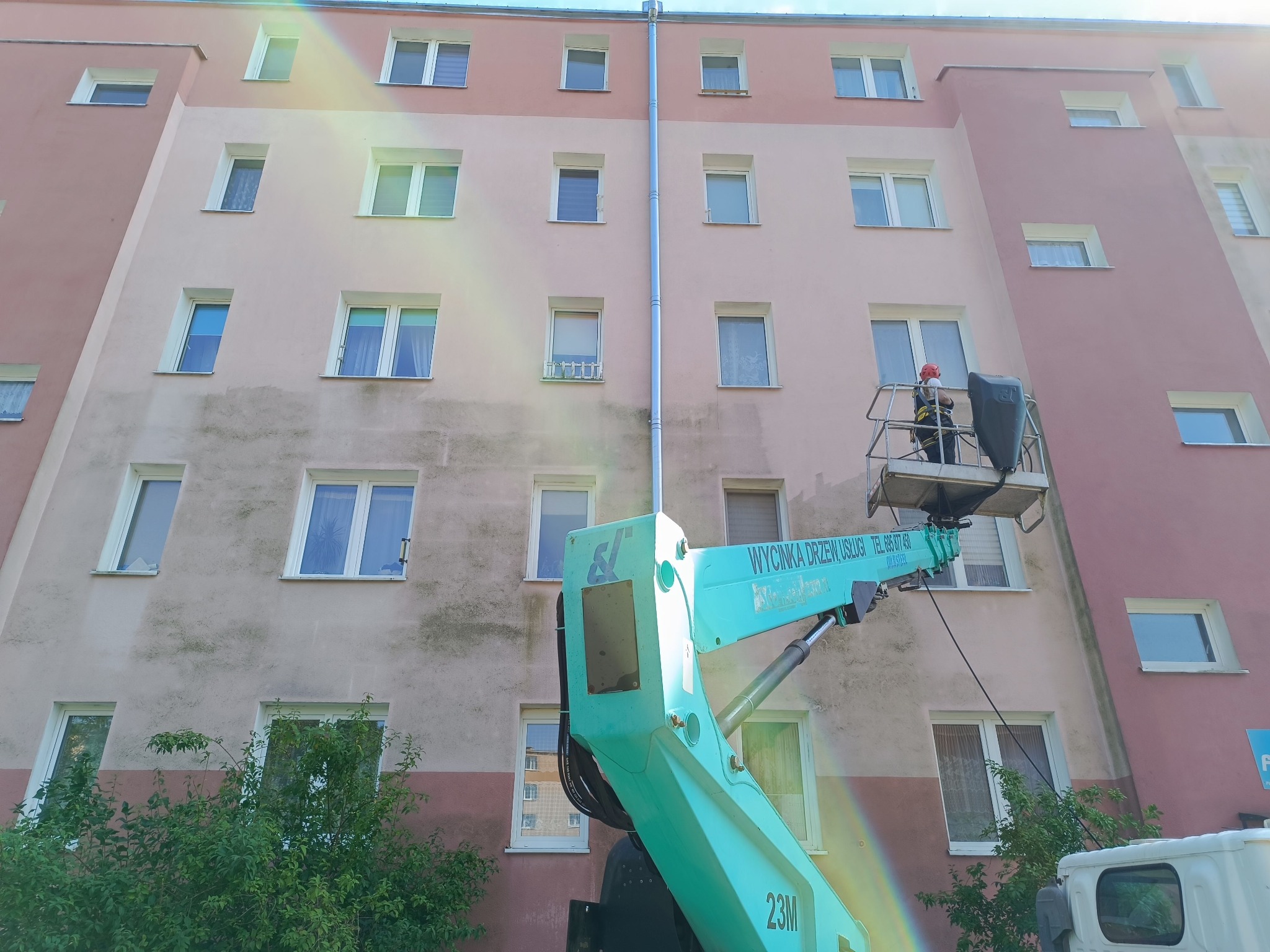 Pracownik w koszu podnośnika czyści rynnę na tle bloku mieszkalnego. Widoczny fragment podnośnika koszowego z napisem 'Wycinka drzew, usługi'.