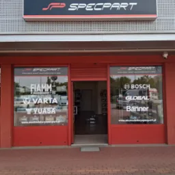 Fasada sklepu motoryzacyjnego Specpart z logo i reklamami akumulatorów marek Bosch, Varta, Yuasa. Czerwone obramowania okien i drzwi, widoczny chodnik z kostki.