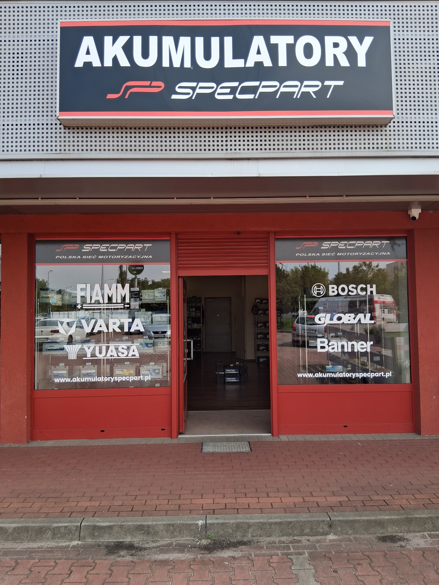 Fasada sklepu motoryzacyjnego Specpart z logo i reklamami akumulatorów marek Bosch, Varta, Yuasa. Czerwone obramowania okien i drzwi, widoczny chodnik z kostki.