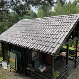 Brązowy dach z blachodachówki na drewnianym domku jednorodzinnym z werandą, otoczonym zielenią i drzewami. Widok z góry.