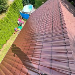 Ekspresowa Naprawa Dachow - Czerwono-brązowy dach z blachy trapezowej w ukośnym ujęciu, cień osoby na dachu, w tle basen i zielone tuje. Widok z góry na nowo położony dach.