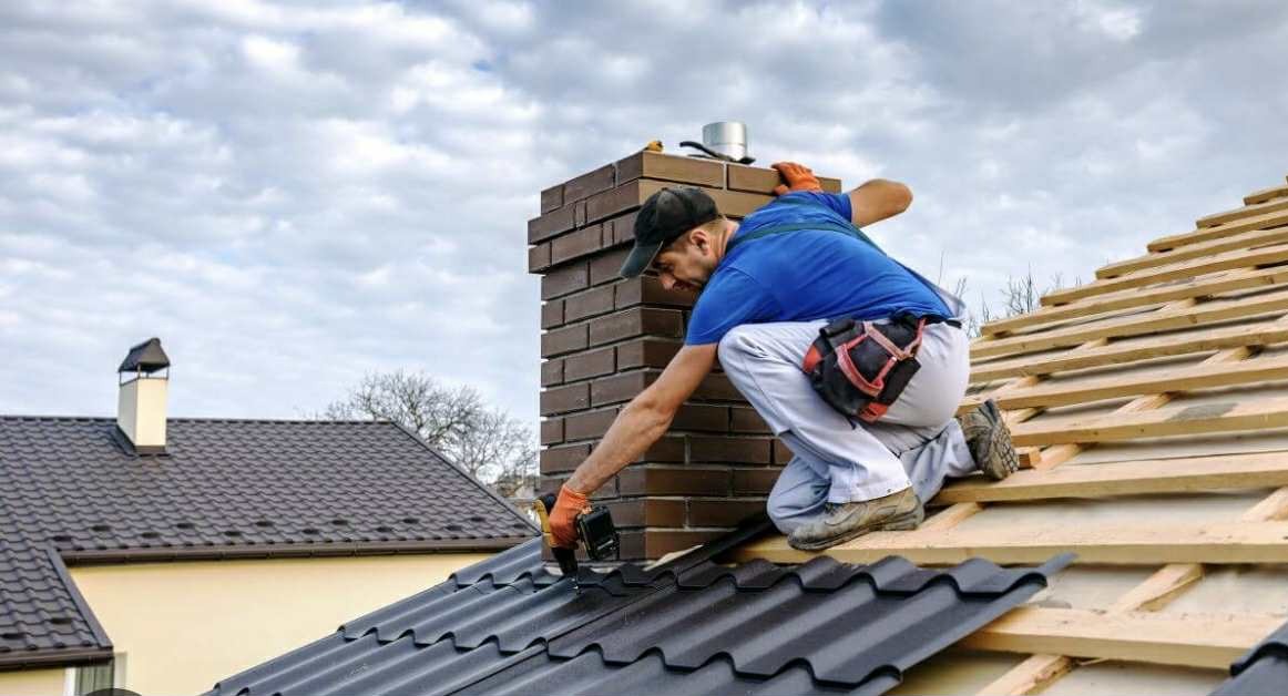 Dekarz montuje ciemną blachodachówkę na drewnianej konstrukcji dachu, obok ceglany komin. Widoczne narzędzia i fragment budynku w tle.