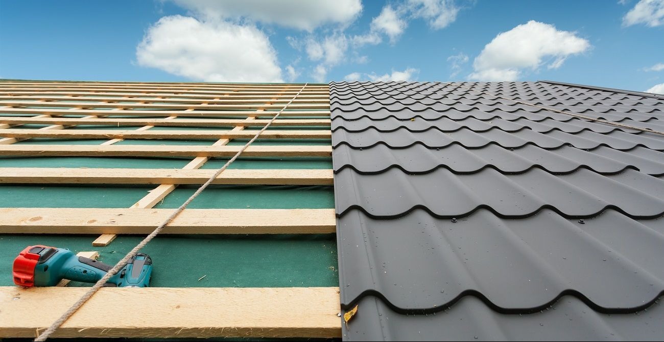 Dach z widoczną konstrukcją drewnianą i częściowo ułożoną blachodachówką w kolorze grafitowym. Na połaci leży wkrętarka. W tle błękitne niebo.