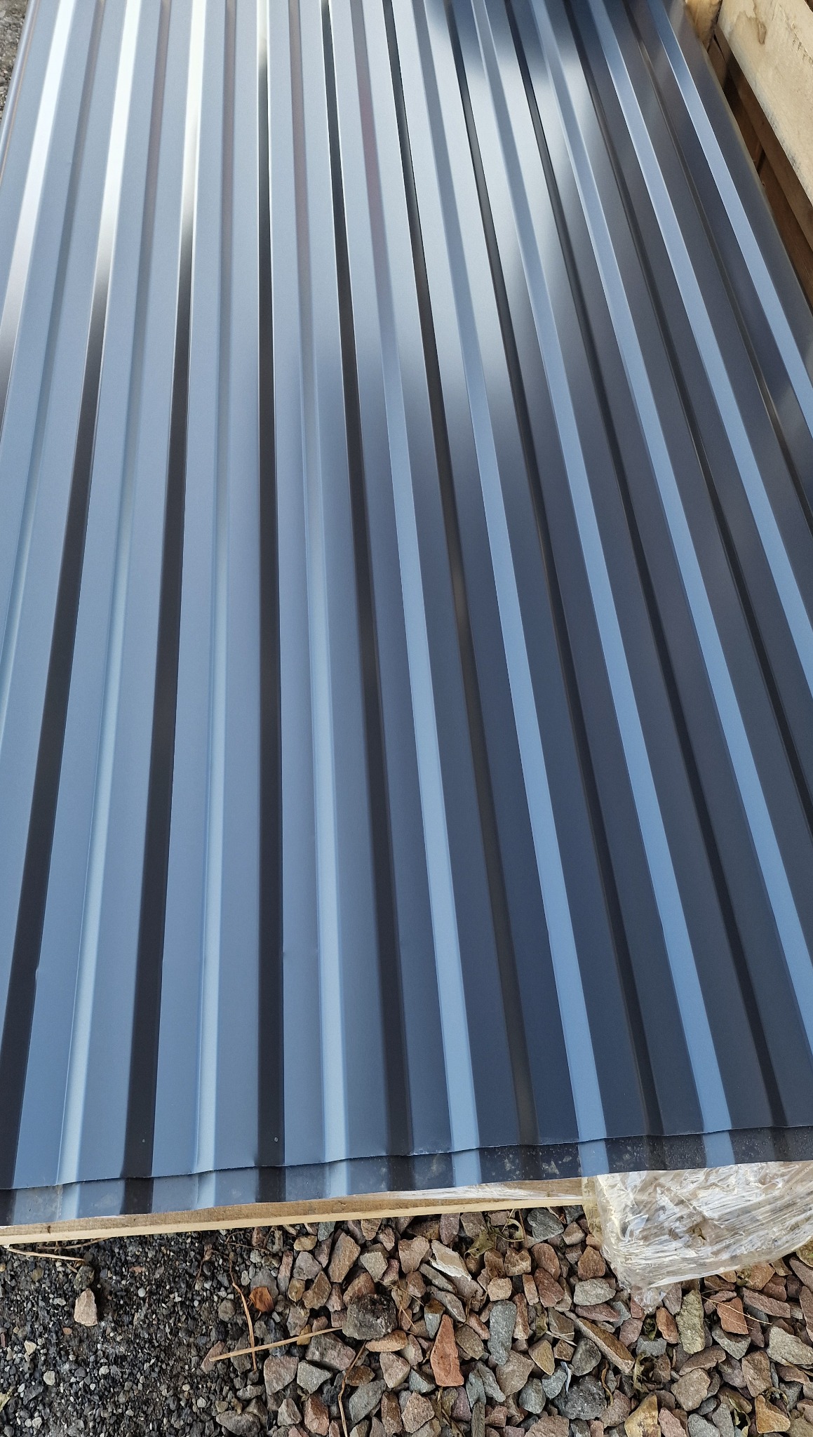 Szare arkusze blachy trapezowej ułożone na palecie, widoczne żwirowe podłoże. Materiał do krycia dachu.