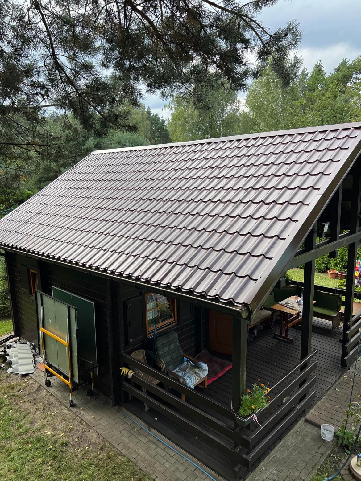 Brązowy dach z blachodachówki na drewnianym domku jednorodzinnym z werandą, otoczonym zielenią i drzewami. Widok z góry.