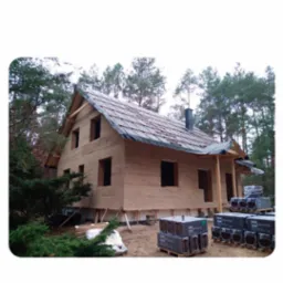 Surowy dom z gliny w lesie na etapie budowy, z widocznym dachem pokrytym folią i paletami z materiałami budowlanymi. Widoczne ściany z gliny.