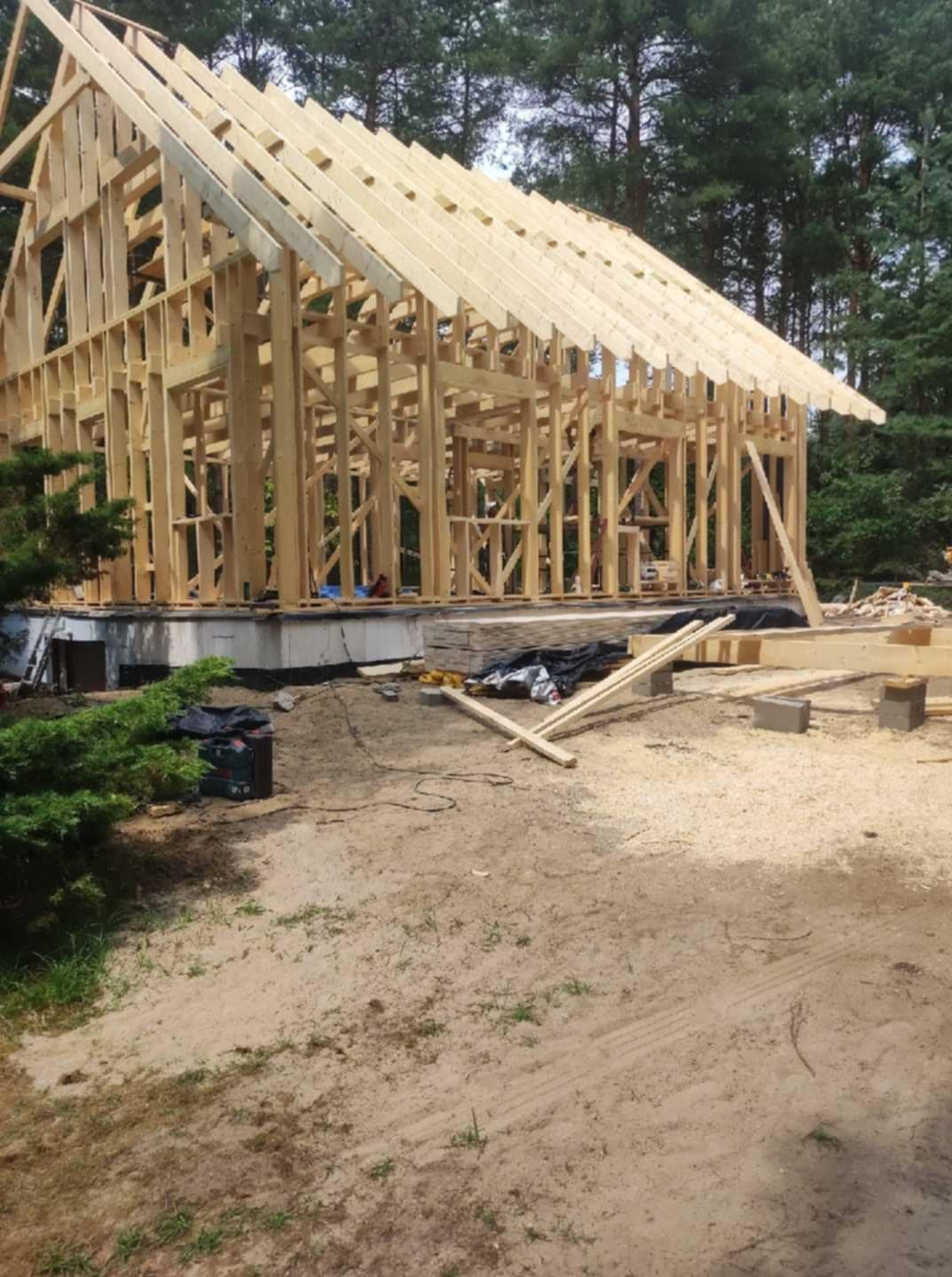 Konstrukcja szkieletowa domu w trakcie budowy, widoczne drewniane elementy i fundament. Otoczenie: drzewa, ziemia. Ujęcie w dzień, z bliska.