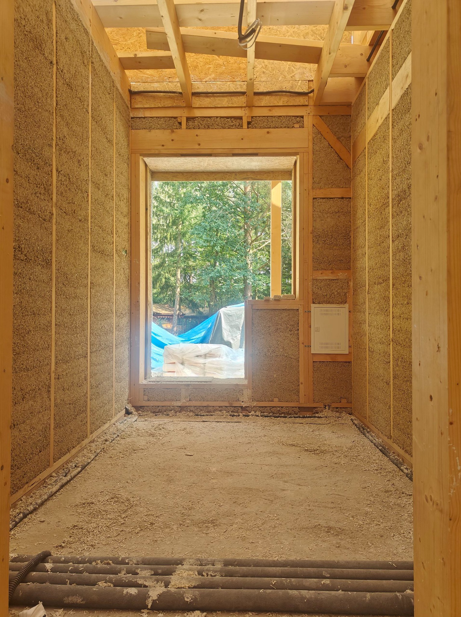 Wnętrze domu szkieletowego w trakcie budowy: ściany wypełnione materiałem izolacyjnym, widoczne drewniane elementy konstrukcyjne i instalacje. Na zewnątrz zieleń.