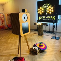 Obrazki z Budki - Drewniana fotobudka na trójnogu z rekwizytami (kapelusze) w pomieszczeniu z dekoracjami urodzinowymi i oświetleniem eventowym. Logo 'Rest-Park' w tle.