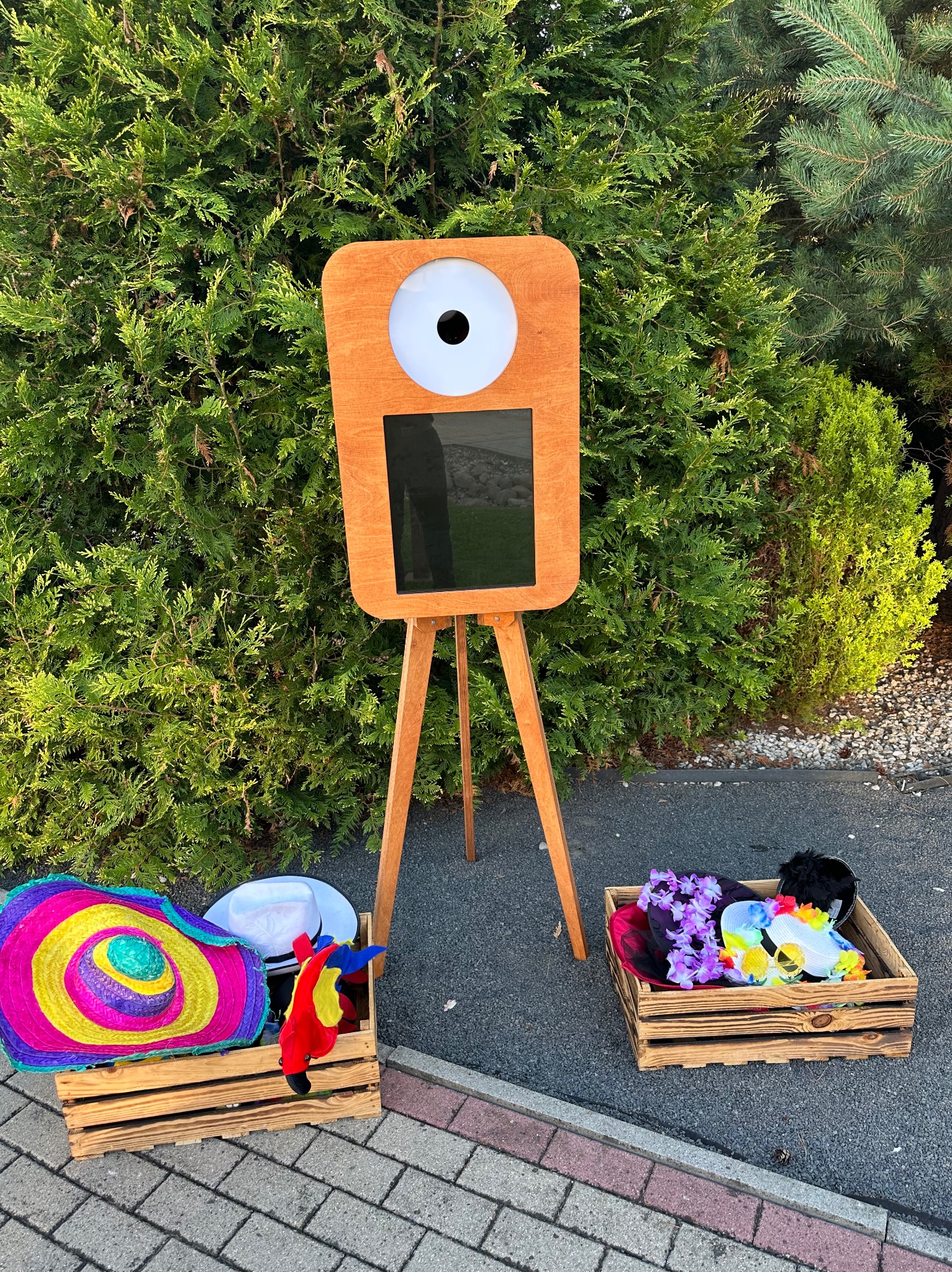 Drewniana fotobudka na trójnogu z rekwizytami (kapelusze, maski) w skrzynkach, ustawiona na zewnątrz, na tle zielonych krzewów. Jasne światło dzienne.