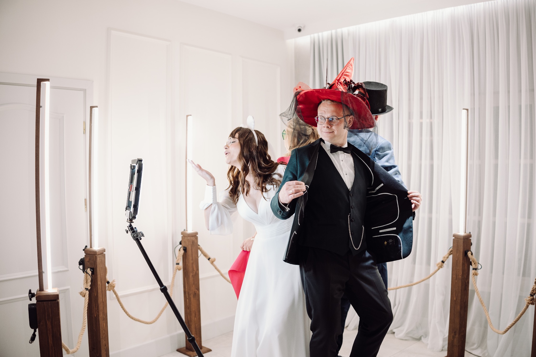 Uczestnicy w przebraniach pozują przed fotobudką z lampami LED i linami w stylu rustykalnym. Mężczyzna w cylindrze i kobieta w sukni ślubnej. Dekoracje imprezowe.