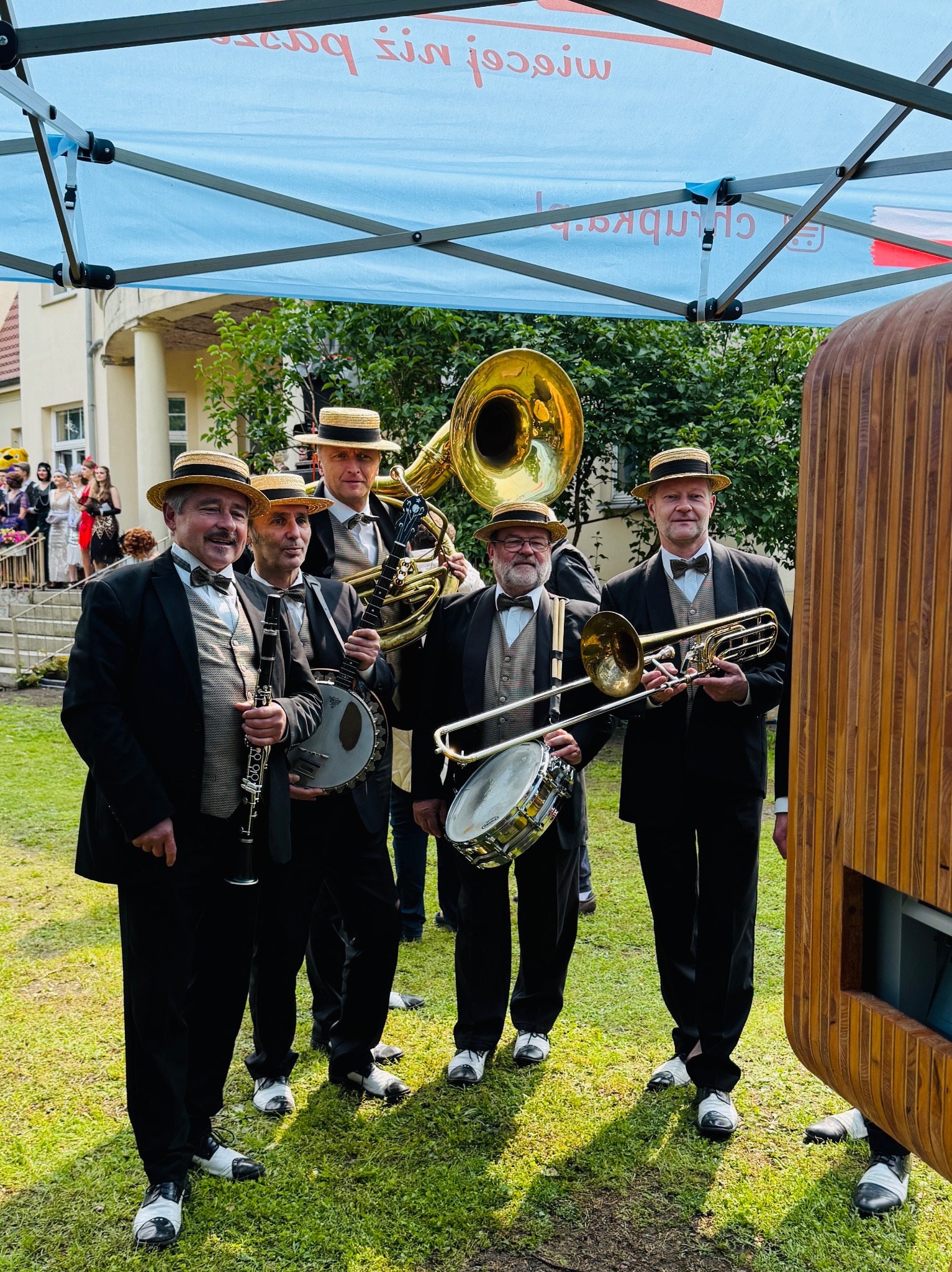 Zespół muzyczny w strojach retro, z instrumentami dętymi i banjo, pozuje na tle budynku i fotobudki podczas plenerowej imprezy.