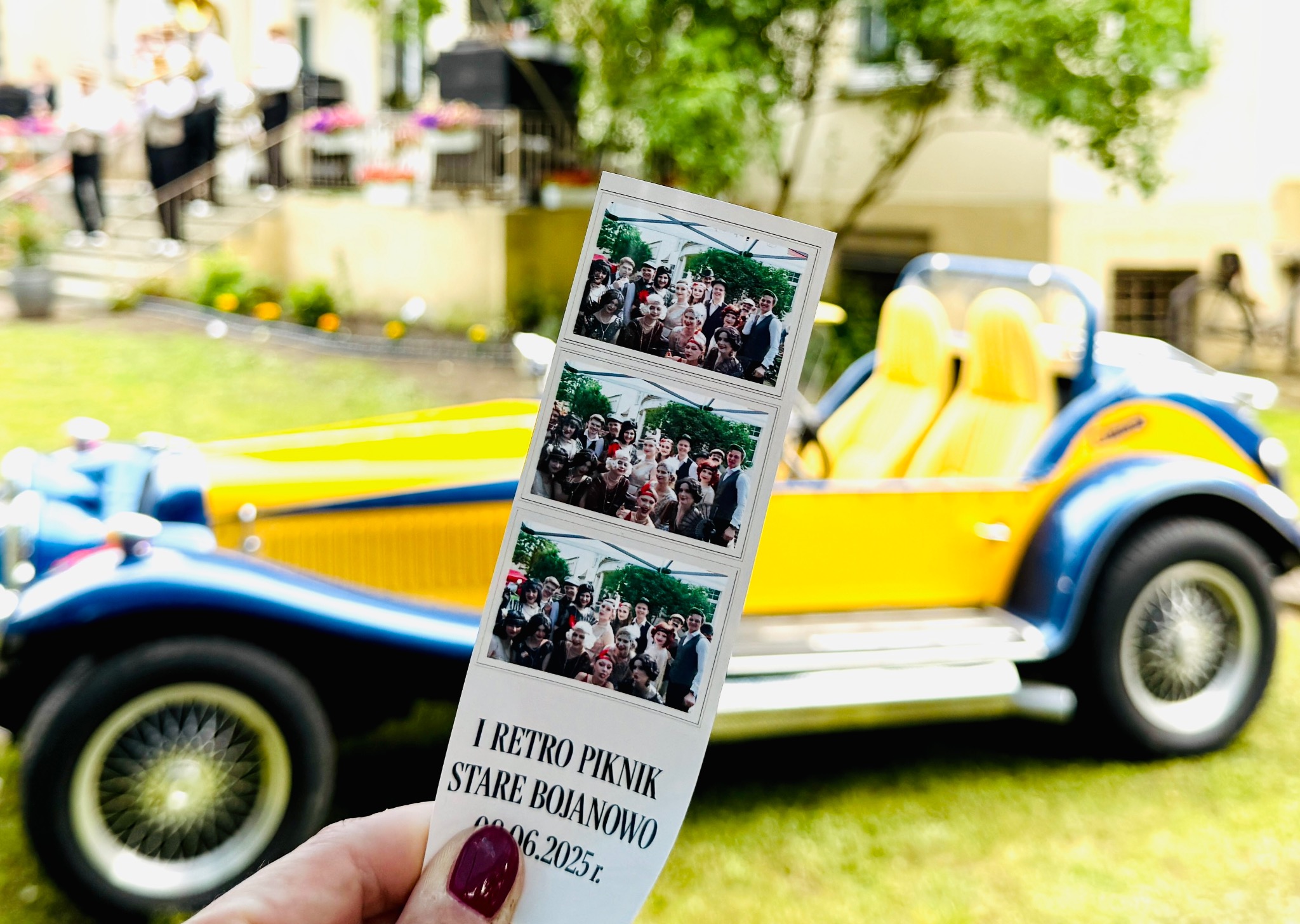 Pamiątkowe zdjęcia z fotobudki z retro pikniku w Kamieńcu, trzymane na tle klasycznego, żółto-niebieskiego samochodu. W tle widoczni muzycy i budynek.