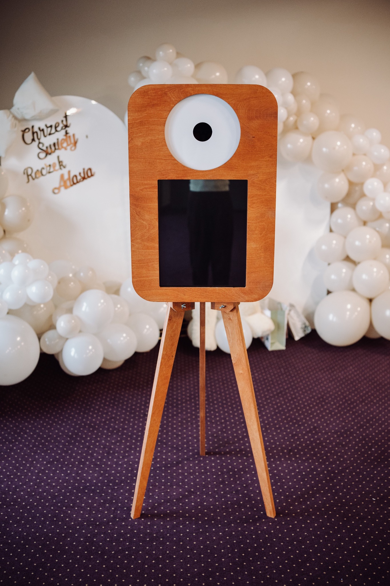 Drewniana fotobudka na trójnogu, ustawiona na tle dekoracji z białych balonów i napisu 'Chrzest Święty'. Widoczny fioletowy dywan w kropki.