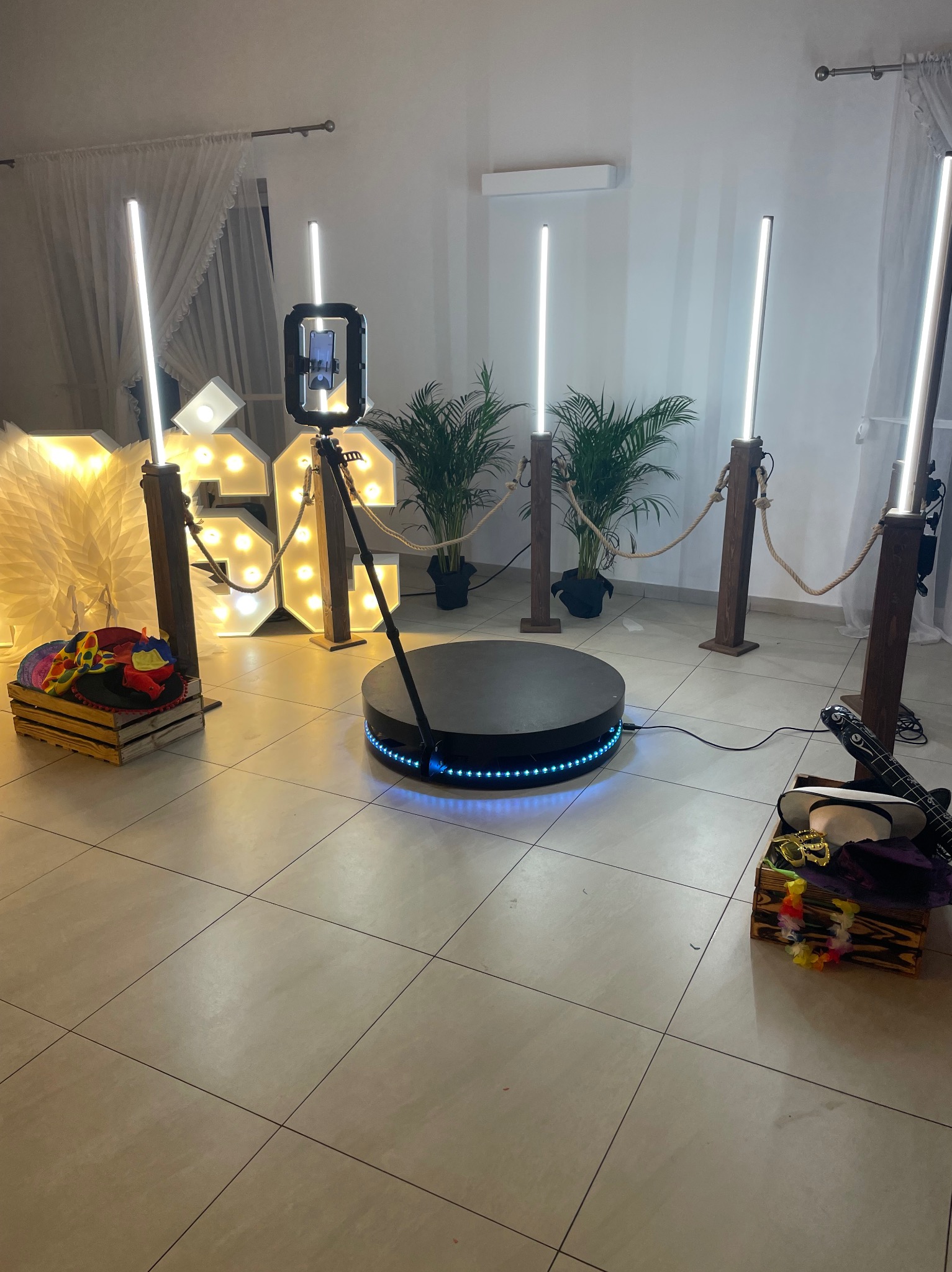 Oświetlona fotobudka 360 z rekwizytami (kapelusze, okulary) na imprezie. W tle dekoracyjne cyfry i rośliny doniczkowe.