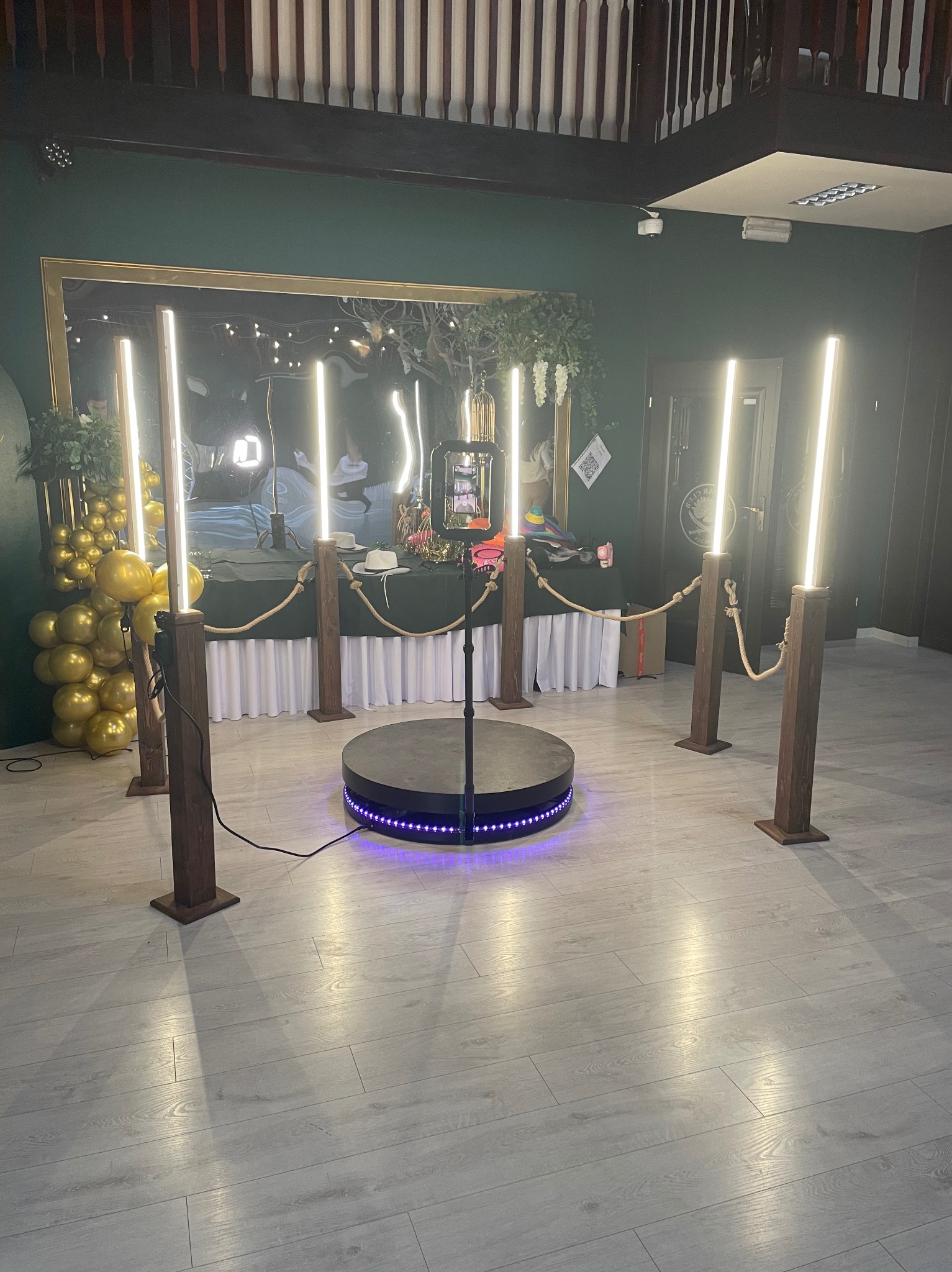Oświetlona platforma 360° z telefonem na statywie, otoczona drewnianymi słupkami z linami, dekoracje z balonów i lustro w tle. Gotowa do użycia fotobudka.