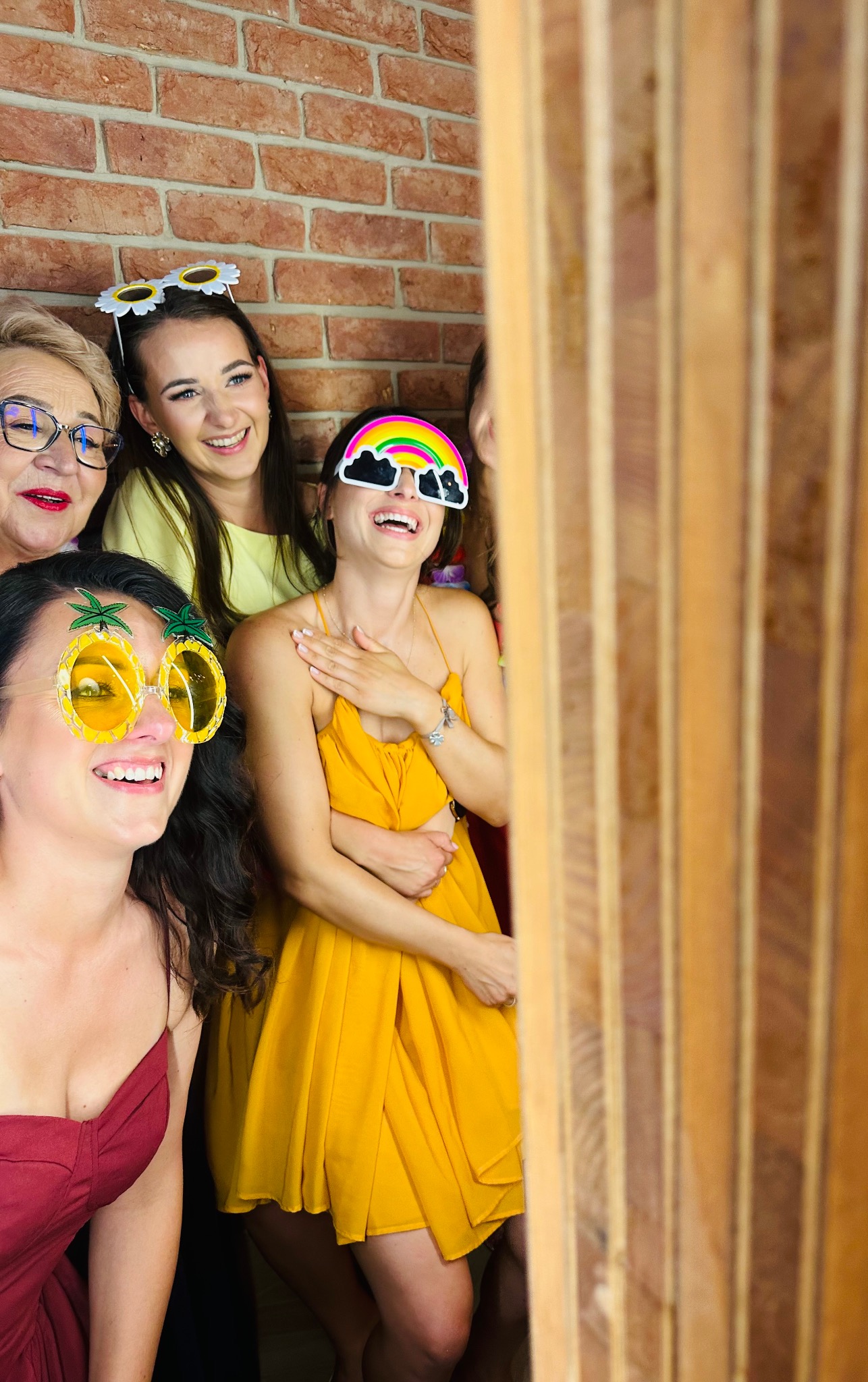 Uśmiechnięte kobiety w zabawnych okularach pozują w fotobudce na tle ceglanej ściany. Radosna atmosfera imprezy.