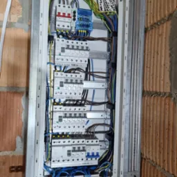 Rozdzielnica elektryczna zamontowana na ścianie z cegły, widoczne bezpieczniki, przewody i oznaczenia. Instalacja w trakcie montażu.