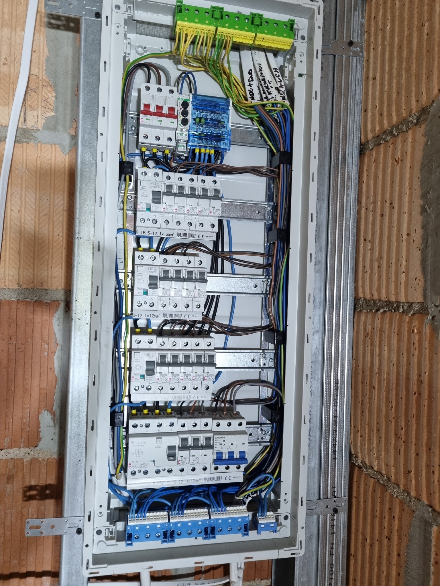 Rozdzielnica elektryczna zamontowana na ścianie z cegły, widoczne bezpieczniki, przewody i oznaczenia. Instalacja w trakcie montażu.