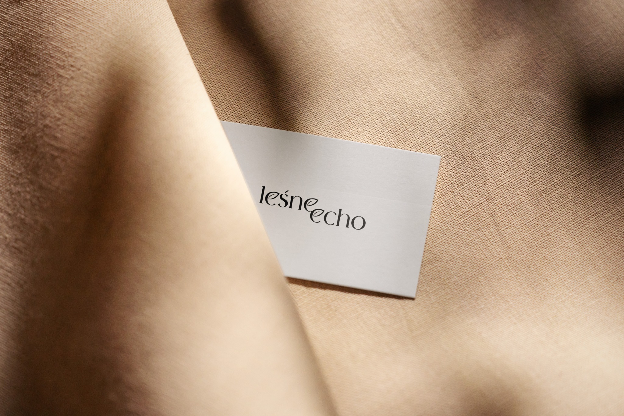 Karta z napisem 'leśne echo' na beżowym tle tkaniny. Minimalistyczny design, elegancki font. Prezentacja logo na materiale.