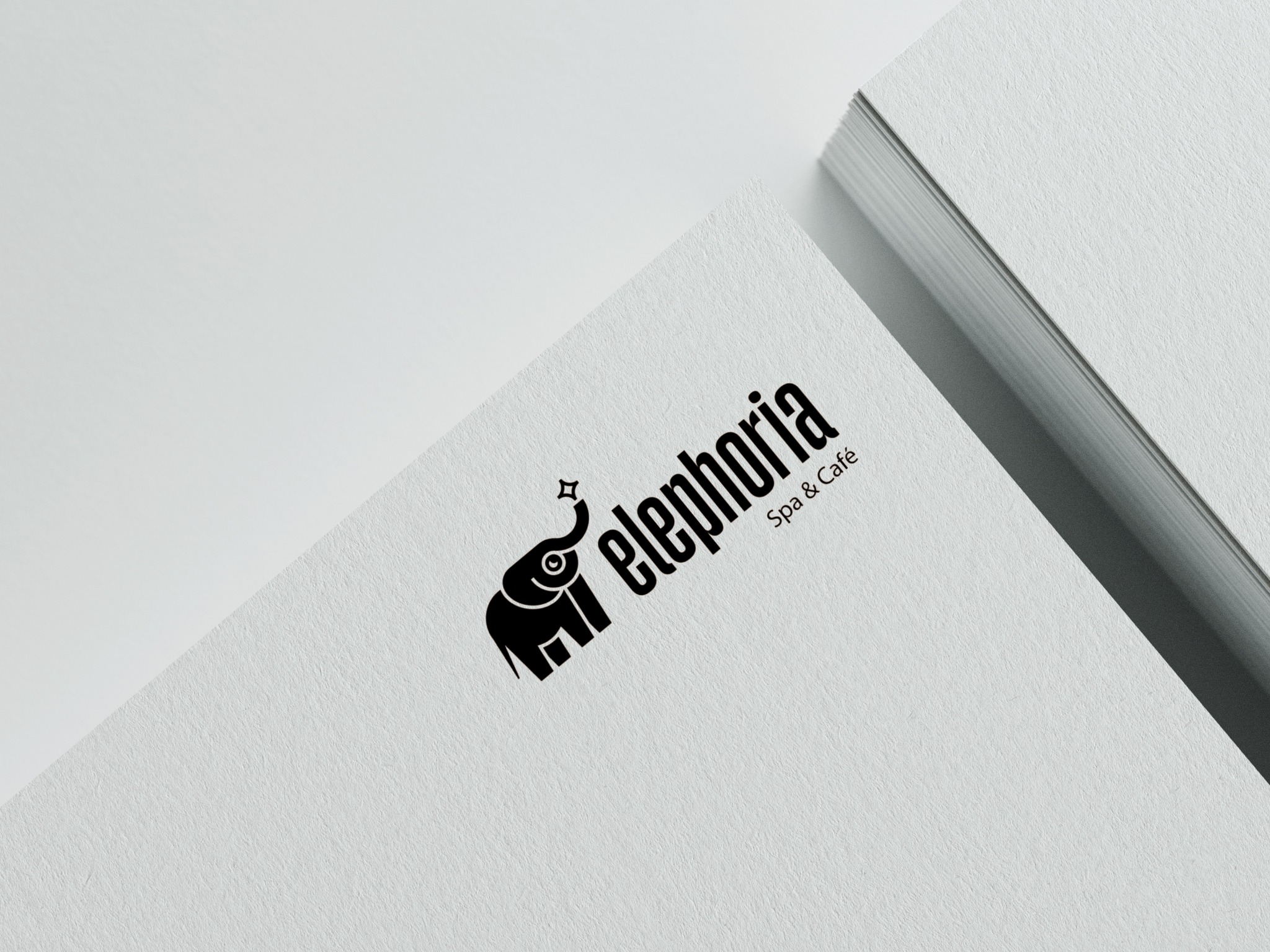 Czarno-białe logo 'Elephoria Spa & Café' z motywem słonia i gwiazdy na wizytówce, ułożone ukośnie na stosie białych kartek. Minimalistyczny design, stonowana kolorystyka.