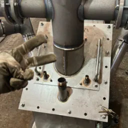 Metalowa konstrukcja z rur i blach, spawana, z widocznymi śladami obróbki. Dłoń w rękawicy pokazuje znak zwycięstwa. Elementy połączone śrubami.