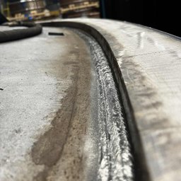 Klaudiusz Brzóska WELD - Detal spoiny na okrągłym elemencie metalowym, widoczne ślady obróbki i wilgoć, surowe oświetlenie przemysłowe, perspektywa z bliska.